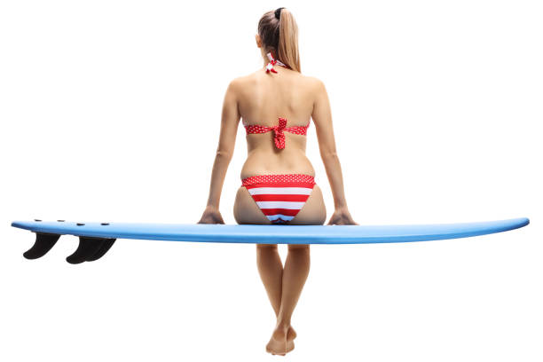 Rear view shot of a young woman in bikini sitting on a surfboard isolated on white background