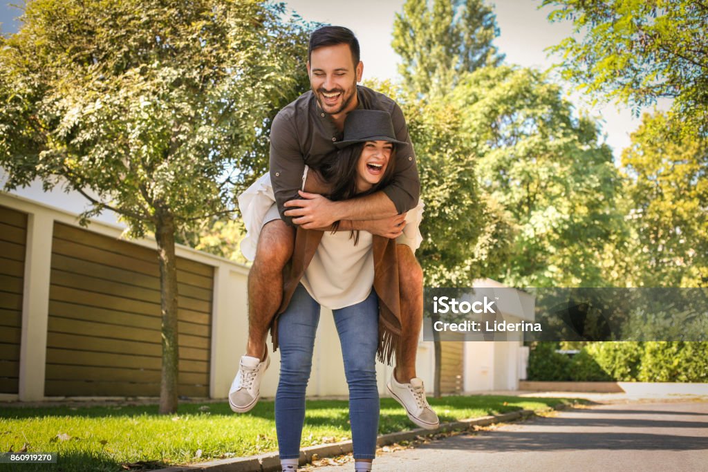 Young couple have fun at city park. Girlfriend carrying his boyfriend on piggyback, they laughing and have fun. Adult Stock Photo