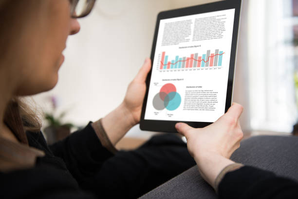 Woman is sitting in a living room looking at a tablet computer showing data, statistics and graphs.