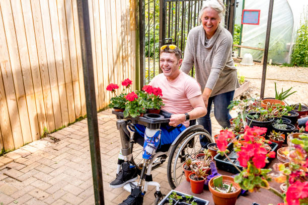 Man Carrying Flowerpots A man in wheelchair carries a tray of flowerpots through the community garden whilst a woman helps him. prosthetic equipment photos stock pictures, royalty-free photos & images