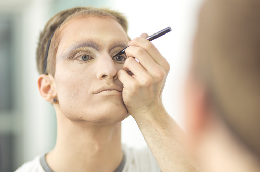 A young man in his 20s does makeup and gets dressed in drag before going out for a night on the town. He is applying eyeliner and looking in the mirror.