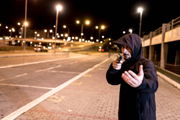 Your money or your life Photo of Robber in black hoodie with a gun holds his hand out for the money, concept of danger, threat and extortion. Man in the dark pointing a firearm at his victim against a night street background, looking at camera man holding gun at night in the city stock pictures, royalty-free photos & images