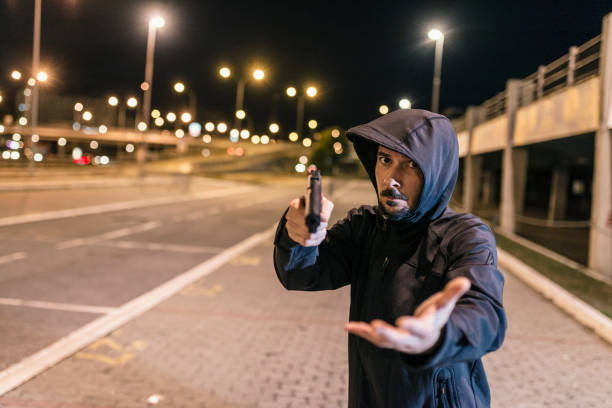 Desperate times call for desperate measures Photo of Robber in black hoodie with a gun holds his hand out for the money, concept of danger, threat and extortion. Man in the dark pointing a firearm at his victim against a night street background, looking at camera man holding gun at night in the city stock pictures, royalty-free photos & images