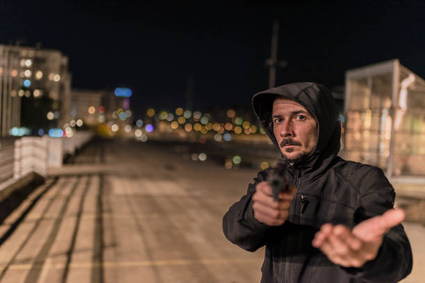 Man with gun pointed at viewer Photo of Robber in black hoodie with a gun holds his hand out for the money, concept of danger, threat and extortion. Man in the dark pointing a firearm at his victim against a night street background, looking at camera man holding gun at night in the city stock pictures, royalty-free photos & images