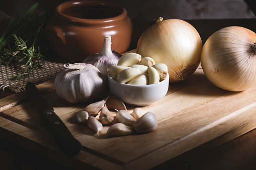 Garlic and onion, near a knife, upon a rustic wooden table, near a cooking pan and some green spices.