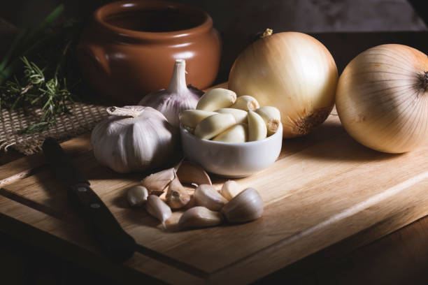 Preparing food Garlic and onion, near a knife, upon a rustic wooden table, near a cooking pan and some green spices. garlic bulb photos stock pictures, royalty-free photos & images