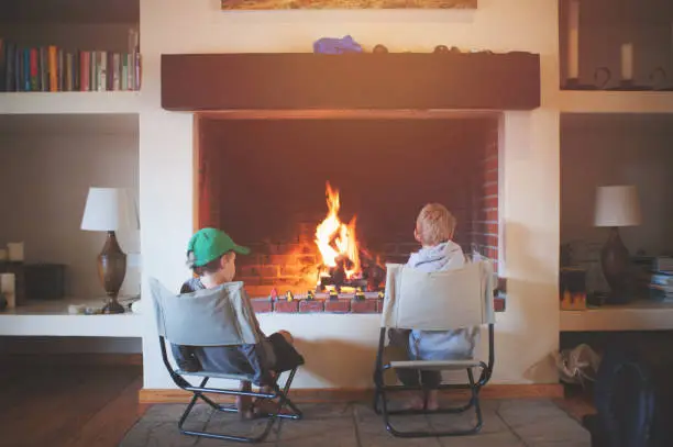 Rear view of two boys sitting on camping chairs at an indoor fireplace with a fire and their play cars in front of them Kromrivier Cederberg Mountains Cape Town South Africa