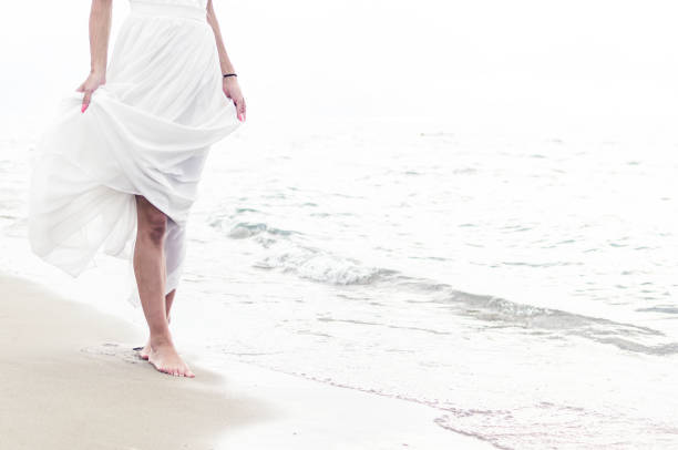 Young woman in white dress walking alone on the sandy beach in the morning. Barefooted legs and foot steps.