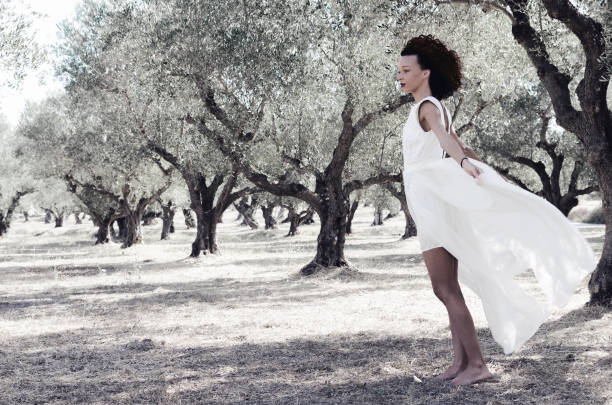 Young beautiful woman in white dress and lust curly hair walking barefooted in olive field.