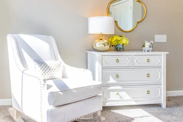 Bright white modern rocking chair in nursery room with chest of drawers, decorations in model staging home, apartment or house