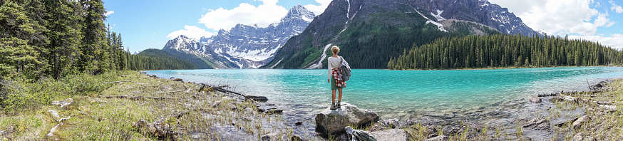 Young woman standing on rock by the lake shore looking at beautiful mountain scenery. Reflection on water surface