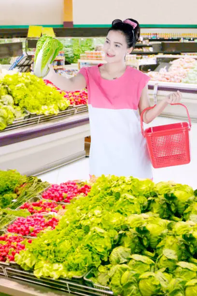 Portrait of pretty woman holding a shopping cart while choosing a fresh vegetable in the supermarket