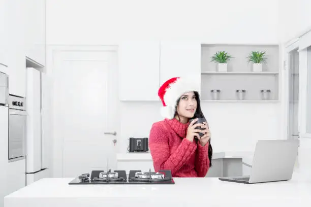 Pretty woman wearing a Santa Claus hat while thinking an idea and sitting with a laptop in the kitchen
