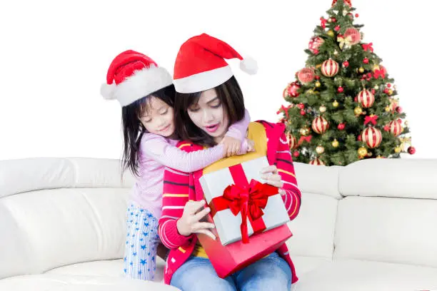 Picture of a pretty woman and her child open a christmas gift while sitting on the sofa and wearing christmas hat with a christmas tree on the background