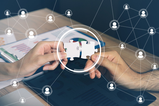 Two hands are holding interlocking puzzle pieces against a backdrop of charts and a digital network graphic, symbolizing connection and collaboration.