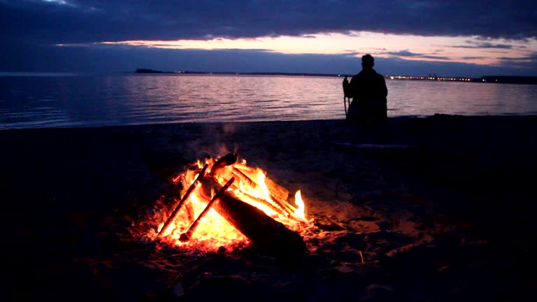 Man sitting by the fire near the river at sunset, Ob River, Siberia,