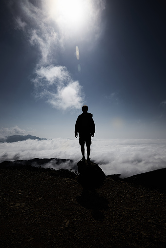 Man silhouette stay on rock peak with clouds below , bright sun and deep blue sky