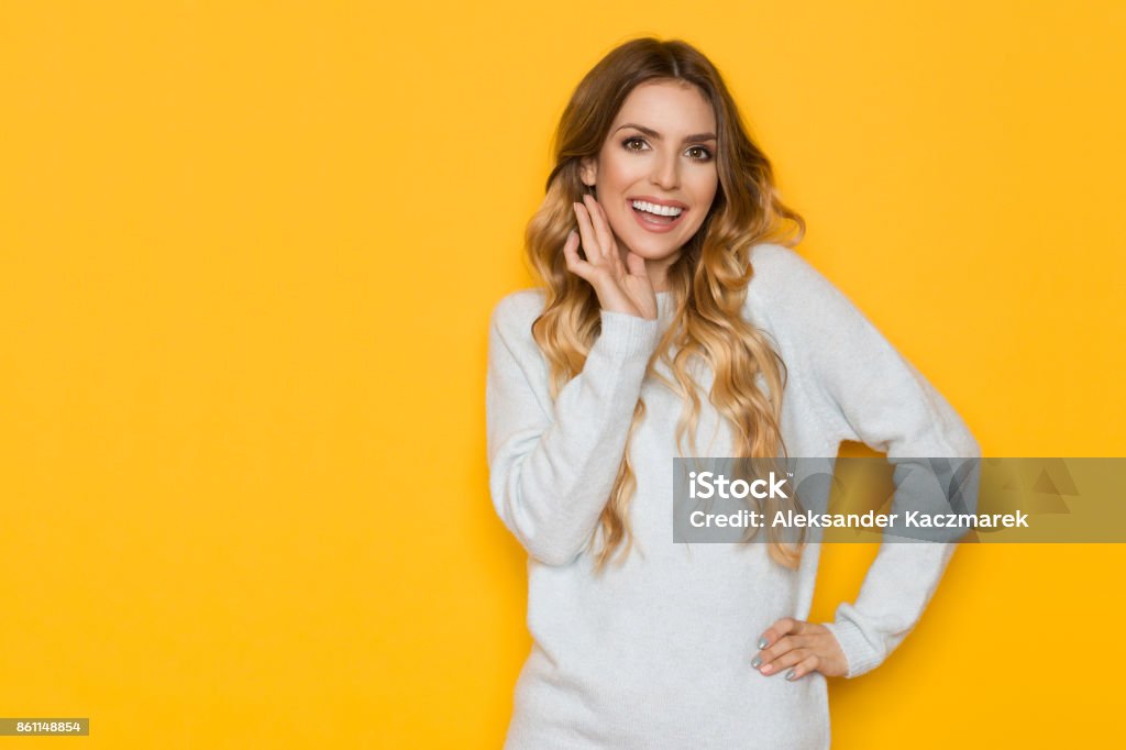 Beautiful Young Woman Is Smiling And Holding Hand On Chin Beautiful young woman in light blue pastel sweater is holding hand on chin, smiling, and looking at camera. Waist up studio shot on yellow background. One Woman Only Stock Photo