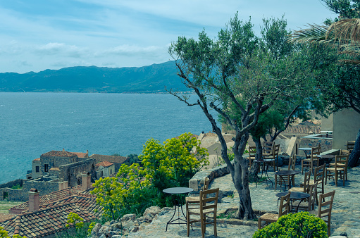 An olive tree surrounded by round iron tables and wooden chairs, typical for traditional greek cafes, overlooking the roofs of houses and the sea in Monemvasia.