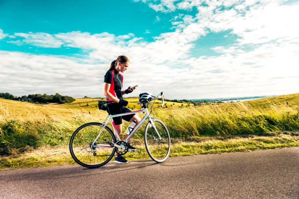 Sporty woman on racing bicycle uses mobile phone while exercising on country road Woman stands next to her racing bicycle on country road. She wears sports clothes and a protective helmet is mounted on the handle bar. She is looking at her smart phone. Maybe she is trying to get a signal, looking for direction, uploading to social media or using an app. super bike stock pictures, royalty-free photos & images