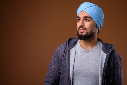 Studio shot of young bearded Indian man wearing casual clothing with blue turban against colored background horizontal shot