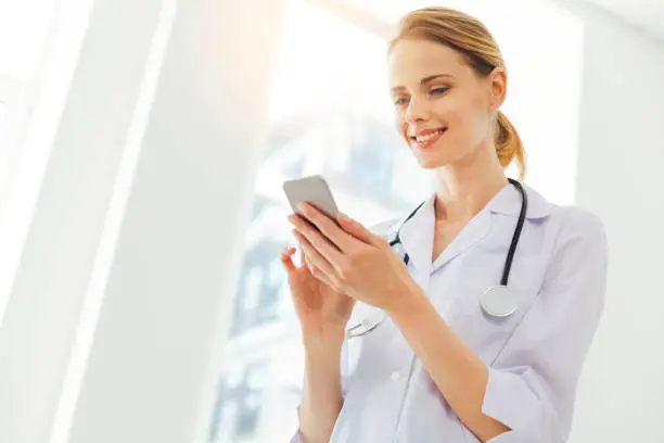 Photo of Joyful young woman in labcoat using smartphone at clinic