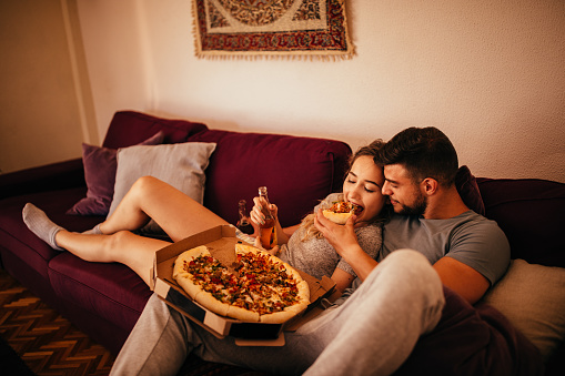 Young hipster couple in love lying on the sofa relaxing, eating pizza and drinking beer