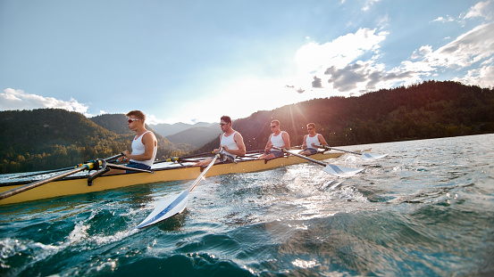 Four male rowers rowing across lake in late afternoon.