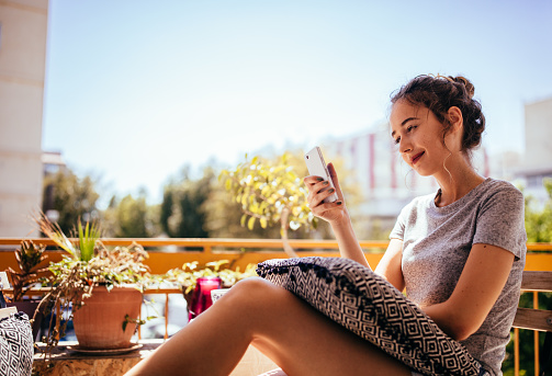 Attractive young woman relaxing on apartment balcony and texting on smartphone