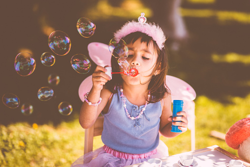 Little Asian girl in fairy costume blowing bubbles at garden tea party with toys