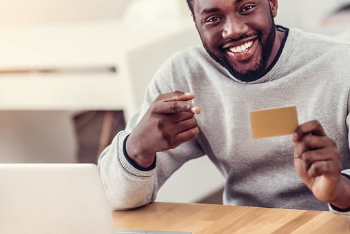 Check this out. Scaled up shot of a millennial African American man grinning broadly while sitting at a table and pointing toward a golden card in his hands.