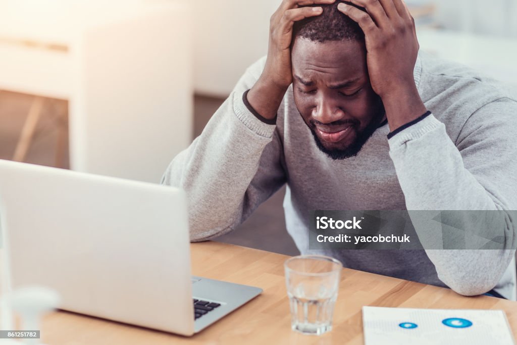 Stressed African American guy crying at work Cannot stand it anymore. Emotionally exhausted young man in casual getting emotional and pressing his head in hands while sitting at a laptop and struggling with a serious project at work. Planning Stock Photo