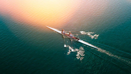 Two female rowers sculling on lake in sunshine.