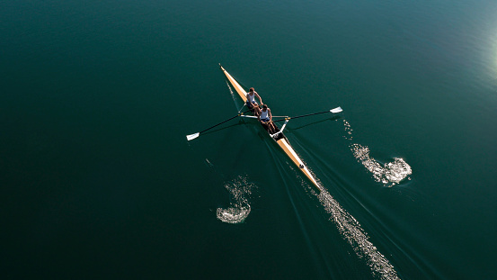 Two male rowers sculling on lake in sunshine.