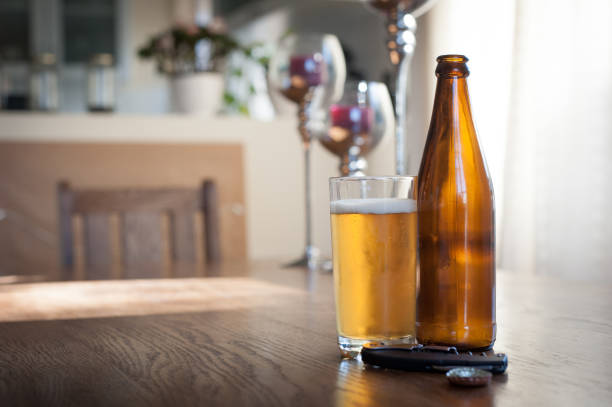 two empty brown glass beer bottles, a glass of beerm bottle top, and a bottle opener on a wooden table within a house