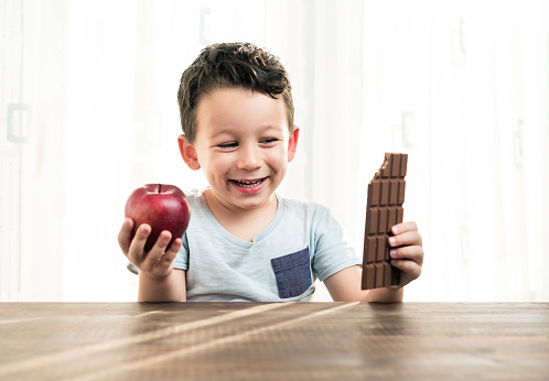 Portrait of child choosing between chocolate and apple. He put an apple and a chocolate on his hands. The child has to make a choice.
