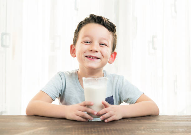 healthy food, drinks, childhood concept - smiling little girl with curly blonde hair dressed in home wear and apron drinking glass of milk over light kitchen background