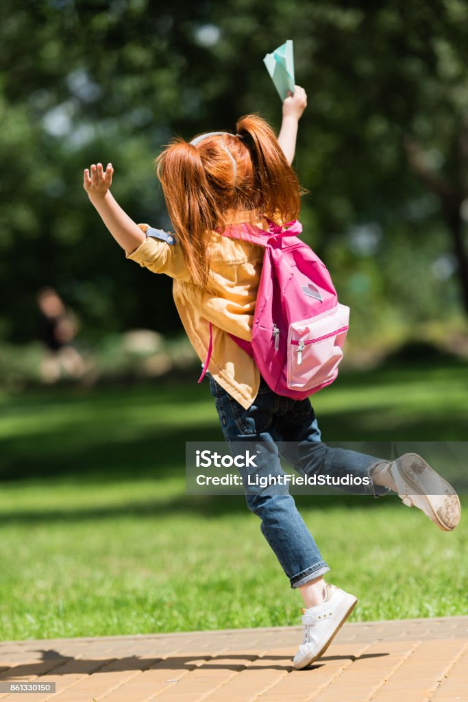 happy redhead schoolchild back view of happy redhead girl with backpack holding paper plane and jumping in park Backpack Stock Photo