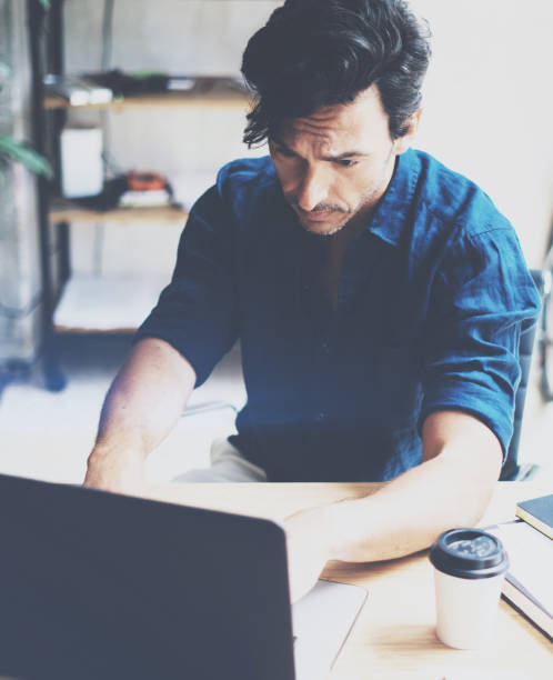 istock Young attractive man working at sunny office on laptop while sitting at wooden table.Businessman analyze digital reports on notebook computer.Blurred background.Vertical. 861332930