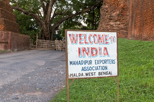 Kotwali gate, 1235-1315. Land border of Bangladesh and India. Welcome to India sign by the right side of the road. Bamboo fence and Indian border guard in the background.