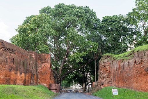 Kotwali gate, 1235-1315. Border of Bangladesh and India. Ancient brick gate with large green trees towering above, a bamboo fence across the road and a welcome to india sign on the right side of the road.