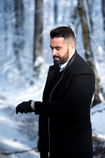 Bearded man in black coat standing outdoors in wintertime and ckecking time on his wristwatch. trees and snow in the blurred background.