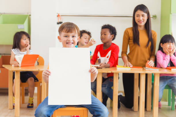 Cute boy holding blank white poster with happy face in kindergarten classroom, kindergarten education concept, Vintage effect style pictures. stock photo