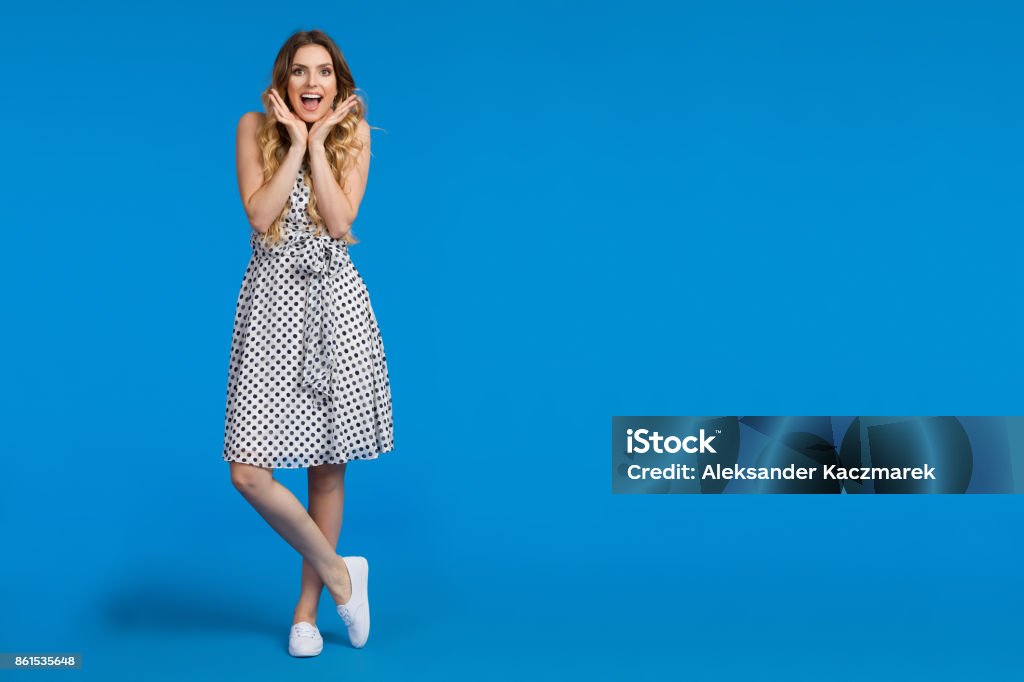 Excited Young Woman On Blue Background Beautiful young woman in white summer dress and sneakers is holding head in hands, shouting and looking at camera. Full length studio shot on blue background. One Woman Only Stock Photo