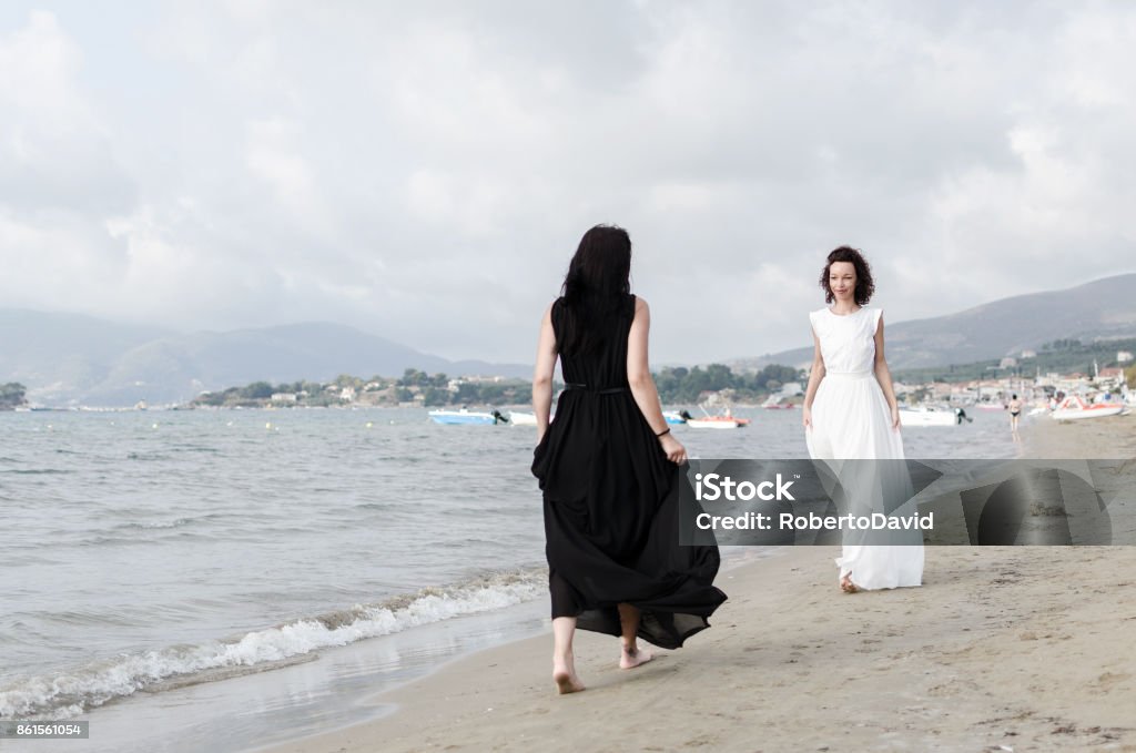 Angel and devil women Angel and devil. Two woman walking in new collection dresses white and black on he sandy beach barefooted. Adult Stock Photo