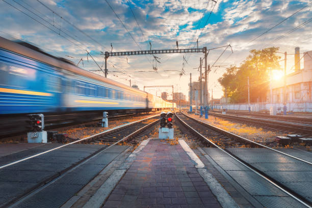 istock Speed passenger train in motion on railroad track at sunset. Railway station with blurred modern commuter train, traffic light, blue sky, clouds and sun. Industrial landscape. Railway platform 861592166