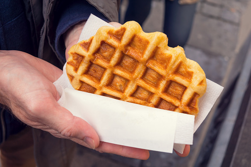 A man's hands are holding a fresh and warm Liege style Belgian waffle wrapped in white paper.