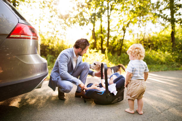 Young father with baby and toddler by the car. Young father with two children by the car. Baby girl in the car seeat. Little boy walking. baby squatting stock pictures, royalty-free photos & images