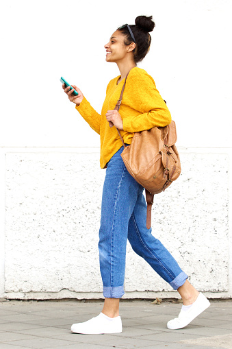 Full length side portrait of stylish young woman walking with bag and mobile phone