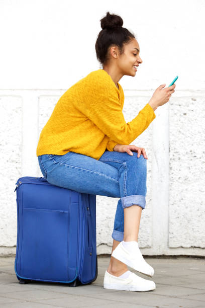 Relaxed female traveler sitting on suitcase and using mobile phone